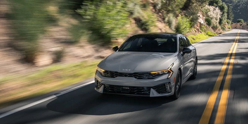Front view of a gray Kia K5 sedan driving on a scenic two-lane road