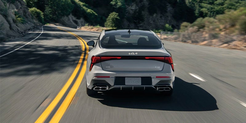 Rear view of a gray Kia K5 sedan driving on a winding road with LED taillights illuminated.